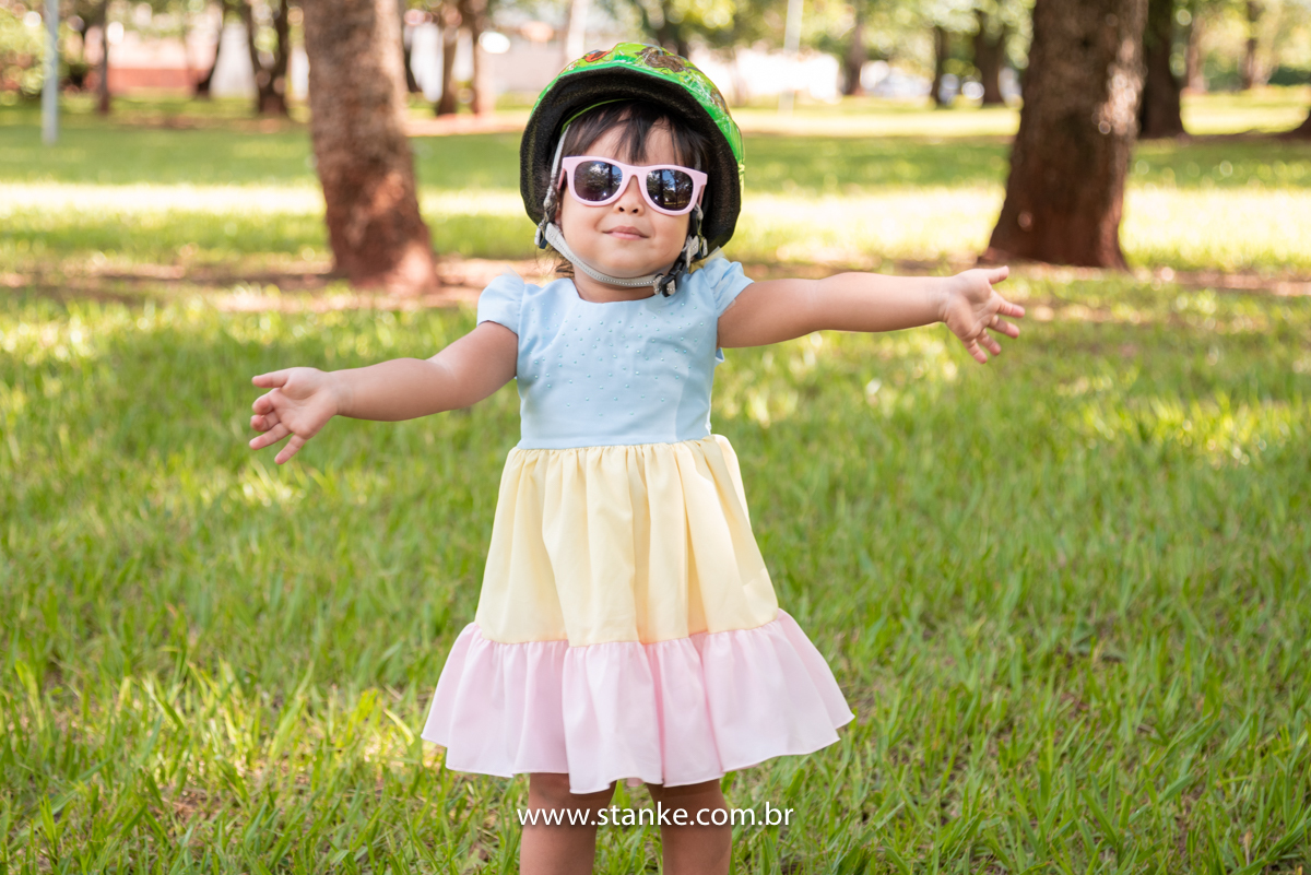 Ensaio infantil da Clara com 2 anos, Aniversariante, com seu vestido colorido e com seus óculos escuros rosa e com capacete de ciclista na cabeça e de braços abertos para o fotógrafo. Fotos feitas no Bosque da Paz, pelo fotógrafo Pedro Stanke. 