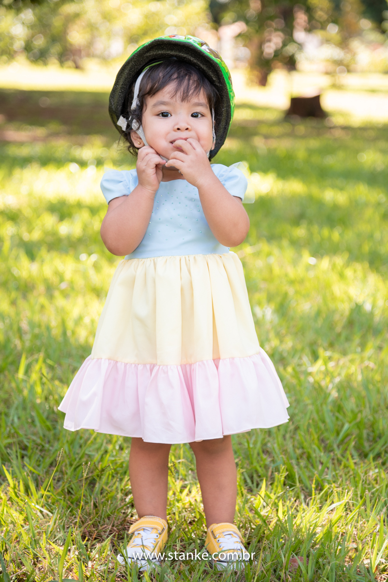 Ensaio infantil da Clara com 2 anos, Aniversariante, de pé, com seu vestido colorido e com capacete de ciclista na cabeça, olhando para o fotógrafo. Fotos feitas no Bosque da Paz, pelo fotógrafo Pedro Stanke. 