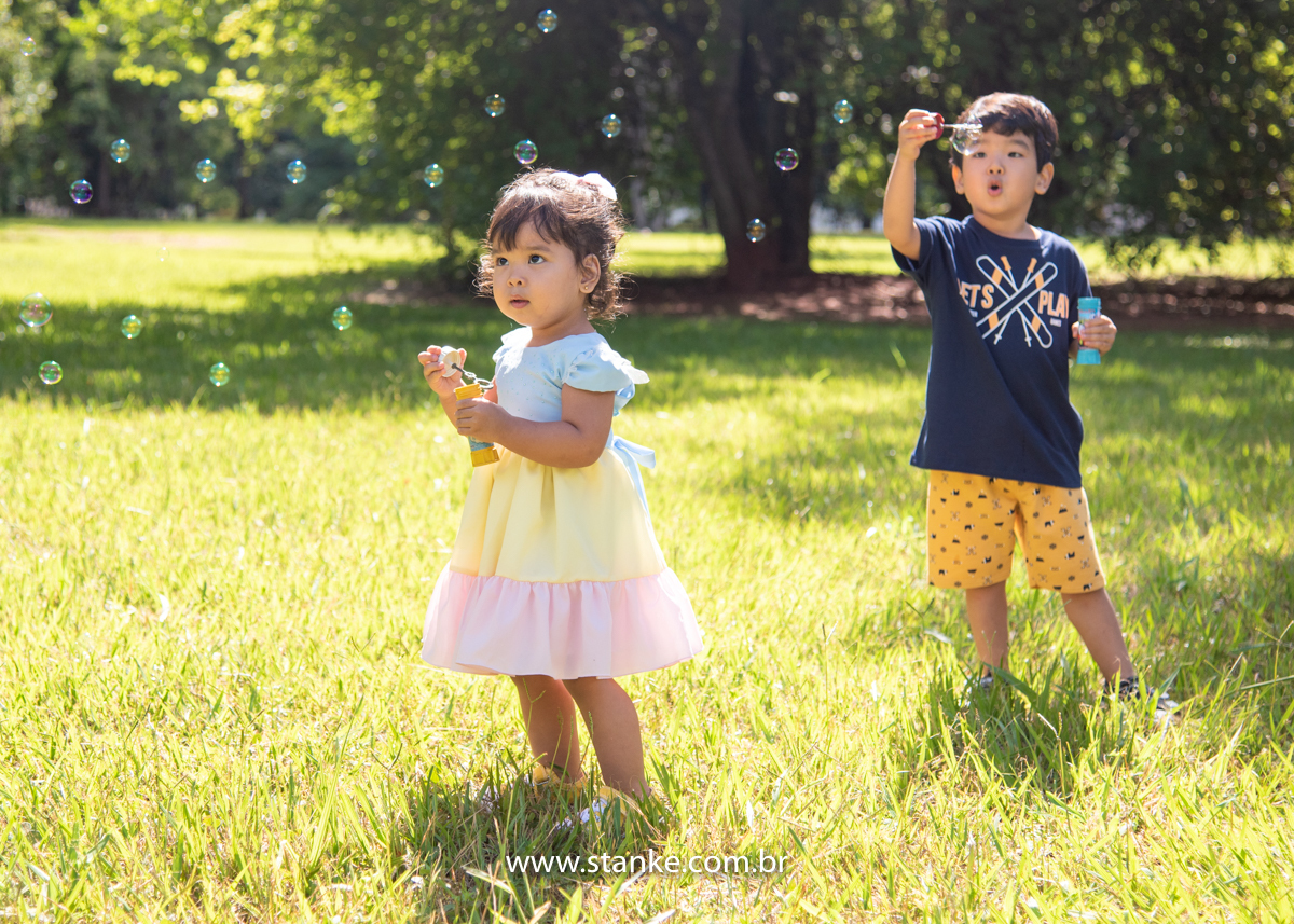 Ensaio infantil da Clara com 2 anos, Aniversariante, com seu vestido colorido e ao fundo seu irmão, ambos fazendo bolhinhas de sabão. Fotos feitas no Bosque da Paz, pelo fotógrafo Pedro Stanke. 