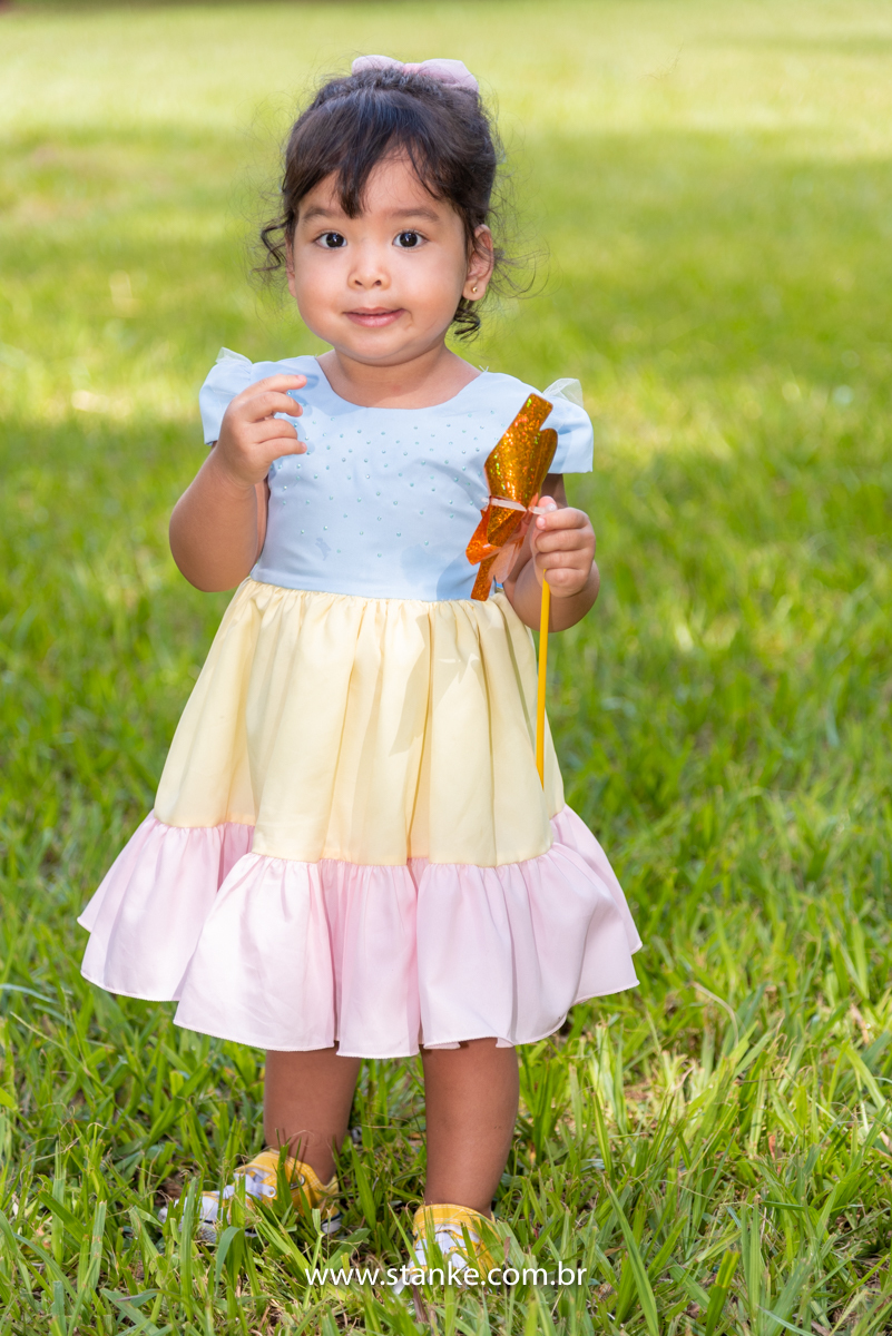 Ensaio infantil da Clara com 2 anos, Aniversariante, com seu vestido colorido, olhando para o fotógrafo e com seu catavento dourado em uma das mãos. Fotos feitas no Bosque da Paz, pelo fotógrafo Pedro Stanke. 