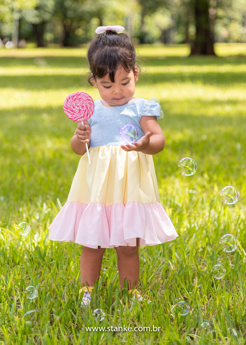 Ensaio infantil da Clara com 2 anos, Aniversariante, com seu vestido colorido,  com seu enorme pirulito em espiral e colorido, e olhando para uma bolha de sabão que caia em uma das suas mãos. Fotos feitas no Bosque da Paz, pelo fotógrafo Pedro Stanke. 