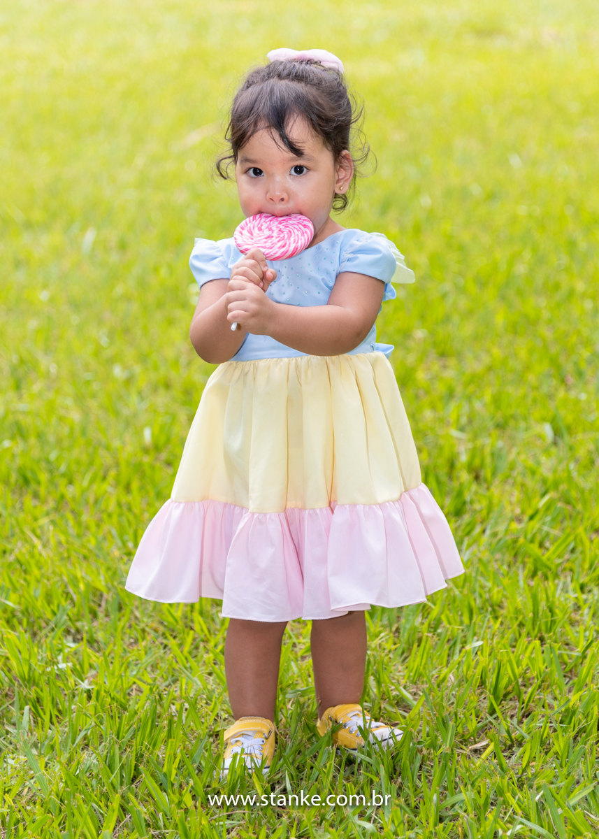Ensaio infantil da Clara com 2 anos, Aniversariante, com seu vestido colorido,  e saboreando o seu enorme pirulito em espiral e colorido e olhando para o fotógrafo. Fotos feitas no Bosque da Paz, pelo fotógrafo Pedro Stanke. 