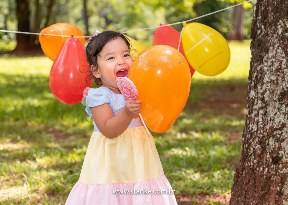 Ensaio infantil da Clara com 2 anos, Aniversariante, com seu vestido colorido,  e saboreando o seu enorme pirulito em espiral e colorido está entre os balões amarrados entre as árvores. Fotos feitas no Bosque da Paz, pelo fotógrafo Pedro Stanke. 