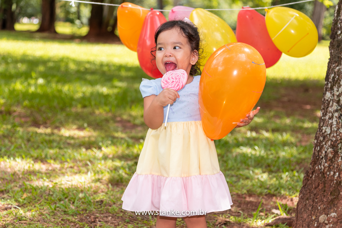 Ensaio infantil da Clara com 2 anos, Aniversariante, com seu vestido colorido,  saboreando o seu enorme pirulito e colorido e está entre os balões amarrados nas árvores e com um deles nas mãos. Fotos feitas no Bosque da Paz, pelo fotógrafo Pedro Stanke. 