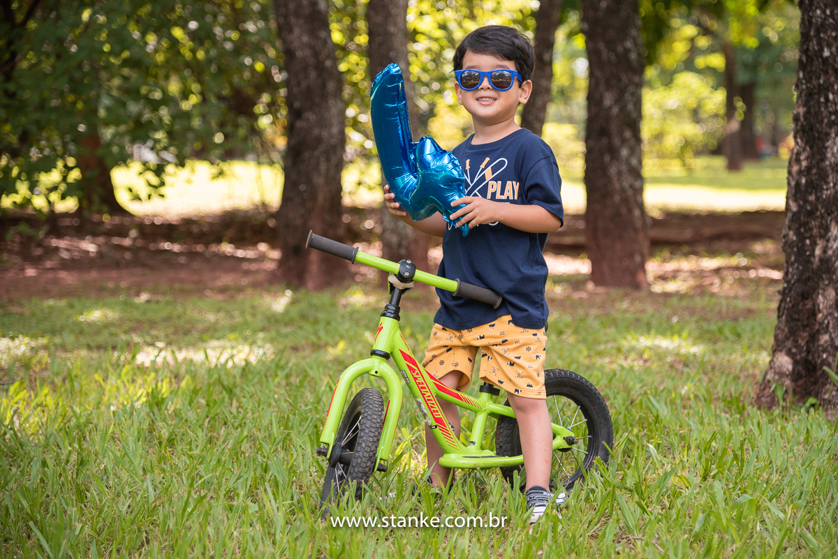 Ensaio infantil do Davi com 4 anos, Aniversariante, com seu com seus óculos escuros azul, sentado em sua bike e segurando, e segurando um balão no formato número 4. Fotos feitas no Bosque da Paz, pelo fotógrafo Pedro Stanke. 
