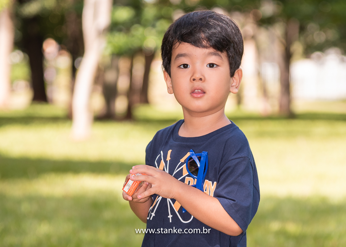 Ensaio infantil do Davi com 4 anos, Aniversariante, sorrindo para câmera enquanto segura o tubinho de bolhinhas de sabão. Fotos feitas no Bosque da Paz, pelo fotógrafo Pedro Stanke. 