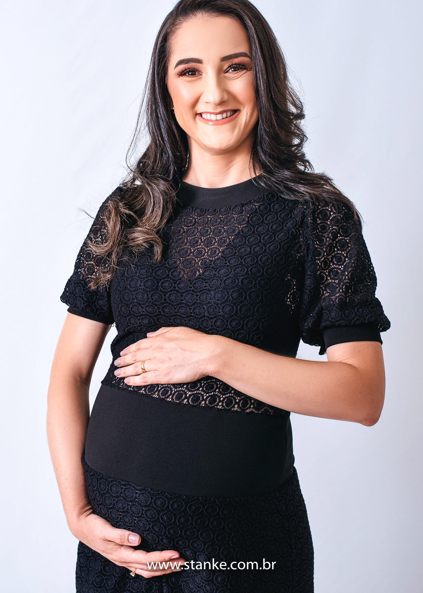 Ensaio gestante em estúdio, no estúdio Stanke Fotografia, gestante com vestido preto todo todo trabalhado e justo, com as mãos na barriga e olhando para câmera, com lindo sorriso. Fotos realizadas pelo fotógrafo Pedro Stanke.
