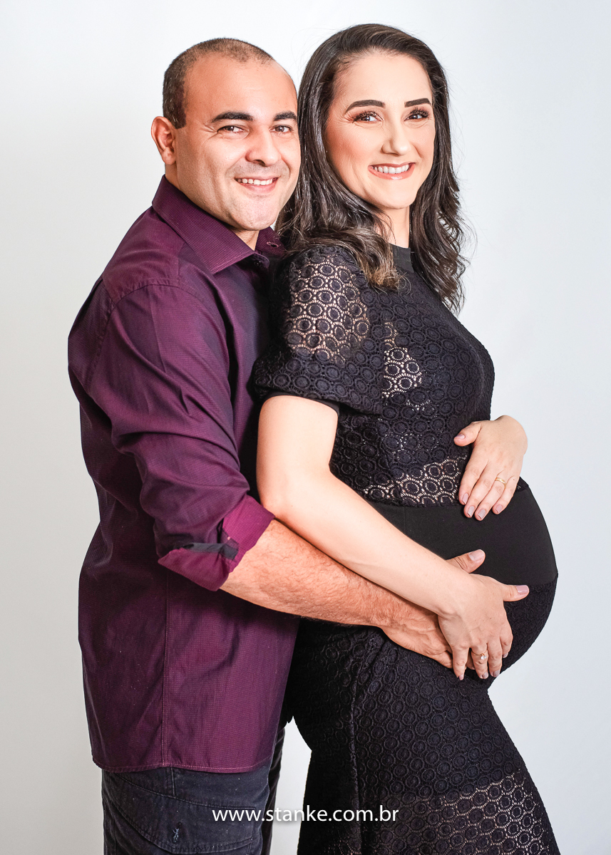 Ensaio gestante em estúdio, no estúdio Stanke Fotografia, gestante com vestido preto todo todo trabalhado e justo, com seu esposo abraçando por traz e olhando para câmera, com lindo sorriso. Fotos realizadas pelo fotógrafo Pedro Stanke.