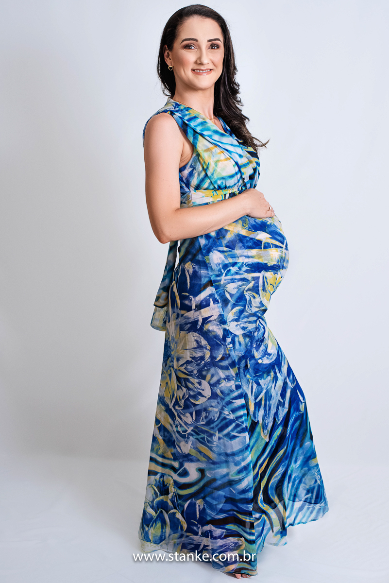 Ensaio gestante em estúdio, no estúdio Stanke Fotografia, gestante com vestido longo e estampado de azul, com as mãos na barriga e olhando para câmera, com lindo sorriso. Fotos realizadas pelo fotógrafo Pedro Stanke.