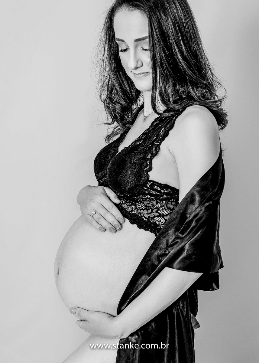 Ensaio gestante em estúdio, no estúdio Stanke Fotografia, uma foto preto e branco da gestante com blusa rendada, lingerie preta e roupão preto caído, com as suas mãos na barriga, e olhando para a barriga. Fotos realizadas pelo fotógrafo Pedro Stanke.