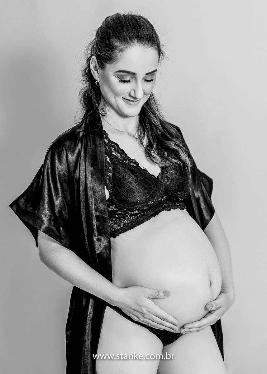 Ensaio gestante em estúdio, no estúdio Stanke Fotografia, uma foto preto e branco da gestante com blusa rendada, lingerie preta e roupão preto com as suas mãos na barriga, e olhando para a barriga. Fotos realizadas pelo fotógrafo Pedro Stanke.