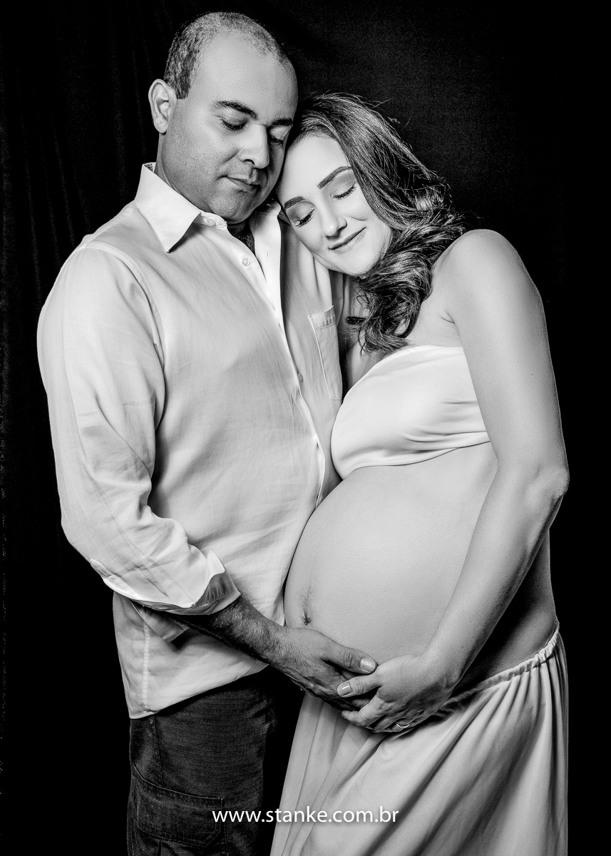 Ensaio gestante em estúdio, no estúdio Stanke Fotografia, foto preto e branco, a gestante com top branco sobre os seios, e saia branca, seu esposo abraçando e as suas mãos na barriga, olhando para a barriga. Fotos realizadas pelo fotógrafo Pedro Stanke.
