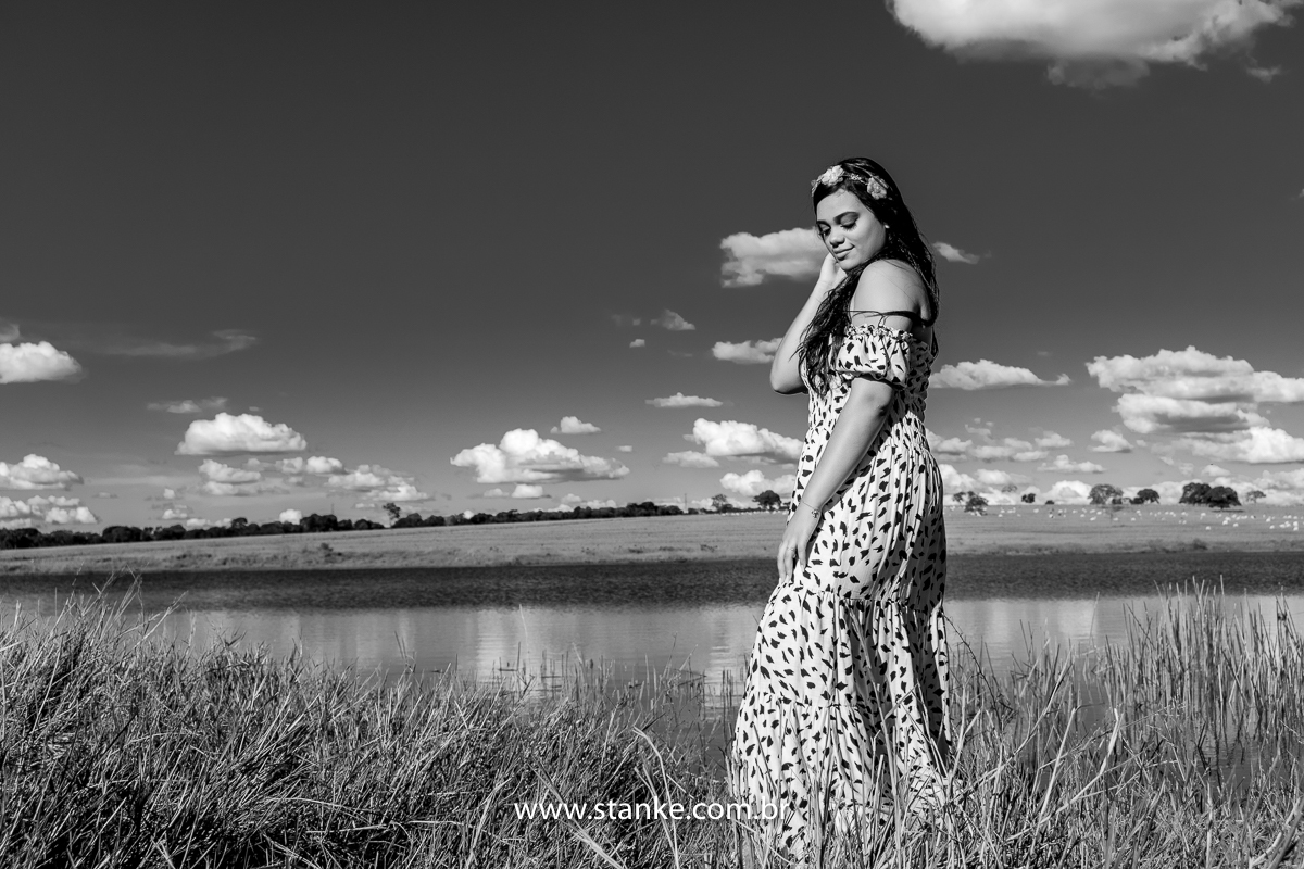 Ensaio 15 anos externo. Preto e branco, 15 anos com vestido longo com manchinhas pretas, e com um tiara de flores na cabeça, olhando para baixo na margem de um a represa. Fotos realizadas Estância Mariana perto Campo Grande-MS. Fotógrafo Pedro Stanke.