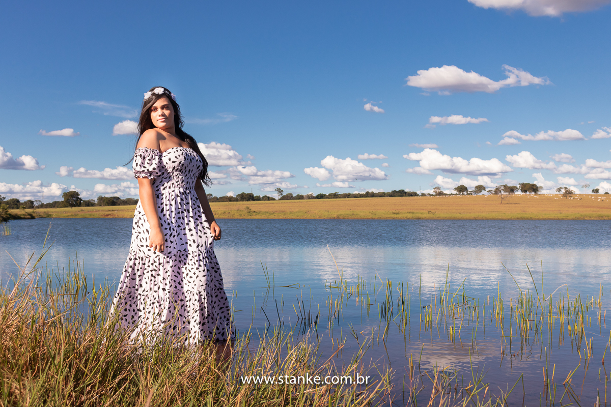 Ensaio 15 anos externo, Aniversariante com vestido longo com manchinhas pretas, tiara de flores, olhando para o lado e com pés na água, na margem de um a represa. Fotos realizadas Estância Mariana perto Campo Grande-MS. Fotógrafo Pedro Stanke.