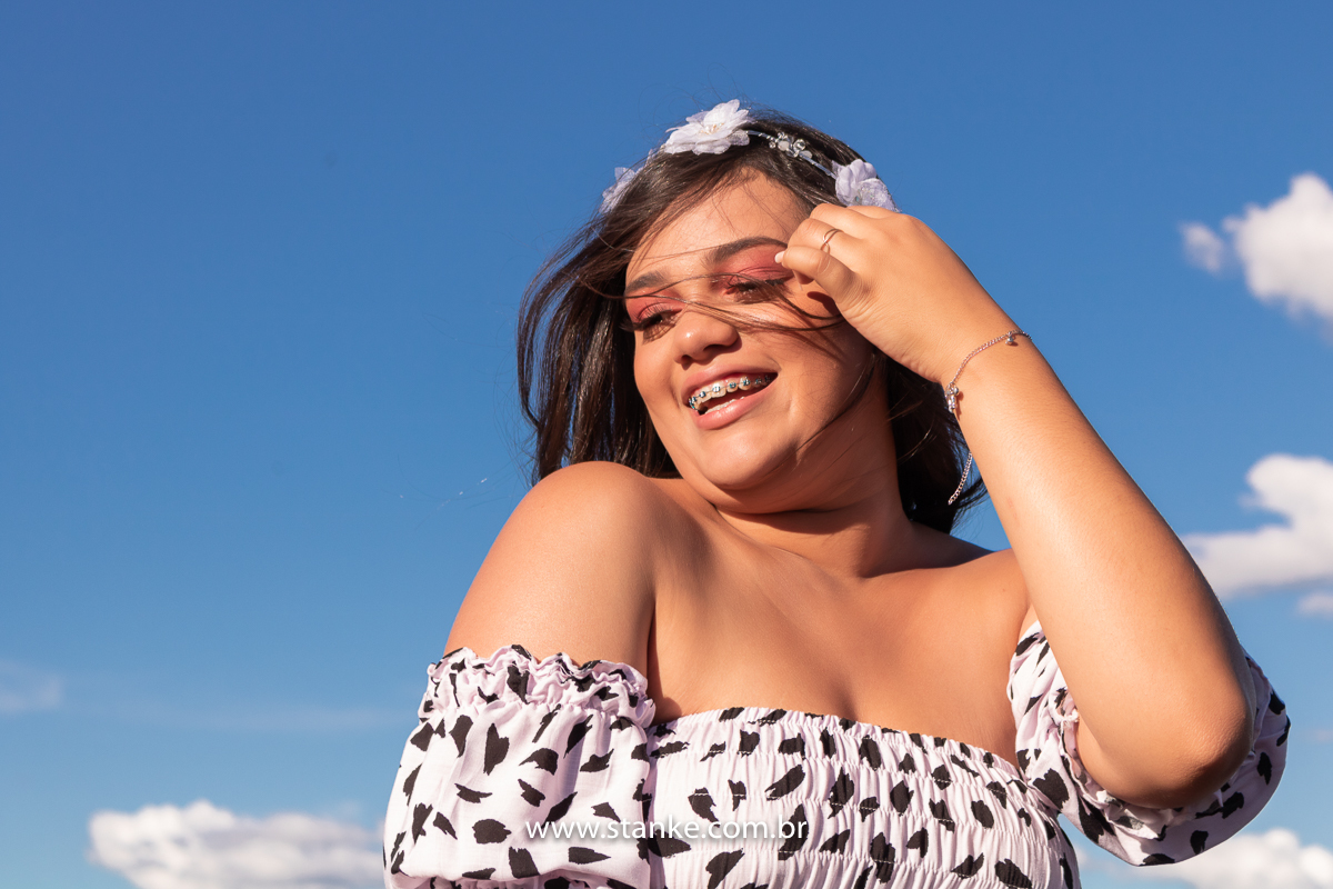 Ensaio 15 anos externo. Aniversariante com vestido longo com manchinhas pretas e tiara de flores, com uma das mãos tirando os cabelos do rosto, com lindo sorriso. Fotos realizadas Estância Mariana perto Campo Grande-MS. Fotógrafo Pedro Stanke.