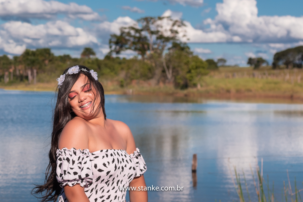 Ensaio 15 anos externo. Aniversariante com vestido longo com manchinhas pretas, tiara de flores, olhando para o lado e com pés na água, e lindo sorriso e cabelos no rosto. Fotos realizadas Estância Mariana perto Campo Grande-MS. Fotógrafo Pedro Stanke.