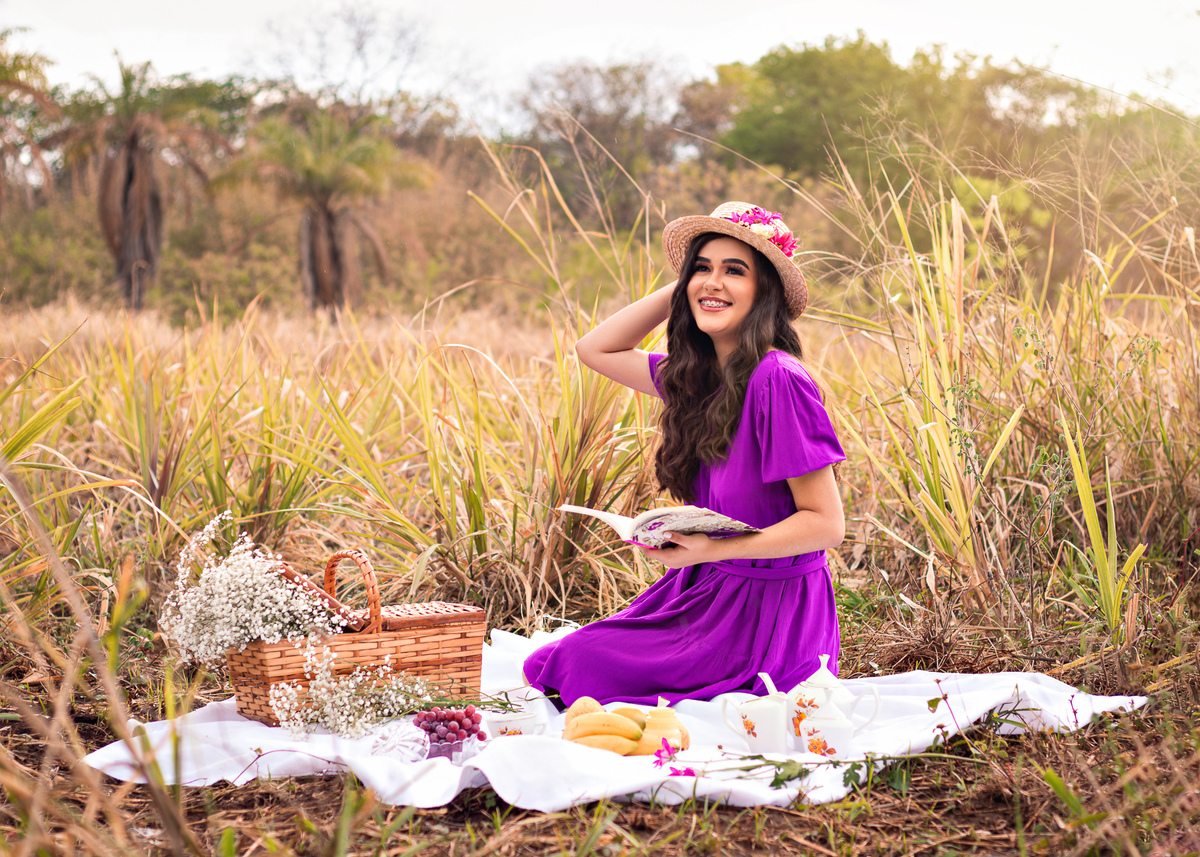 Menina com vestido roxo segurando chapéu e sorrindo olhando o por do sol