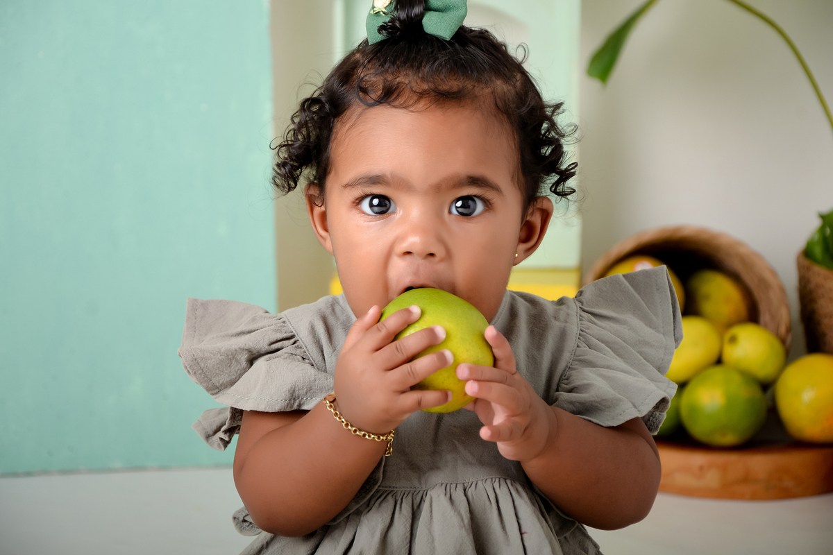 drica studio - ensaio fotográfico infantil - xinguara - frutinha -menina com laranja na boca - cesto com laranja - fundo verde 