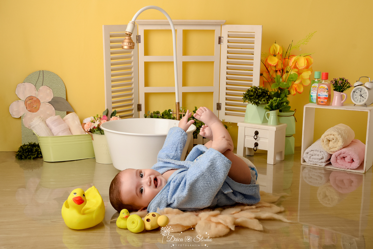 drica studio - ensaio fotográfico - xinguara - bebê deitado no chão -  cenário com fundo amarelo - banheira branca - janela branca - toalhas para compor cenário - flores amarela - patinhos amarelos - bebê vestindo roupão azul 