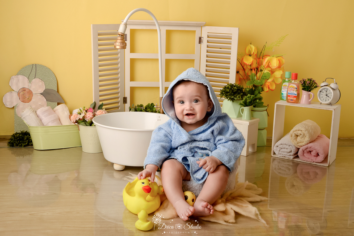 drica studio - ensaio fotográfico - xinguara - menino de roupão com capuz azul claro, brincando com patinhos amarelos - banheira branca - janelinha branca e nicho branco com toalhas dentro