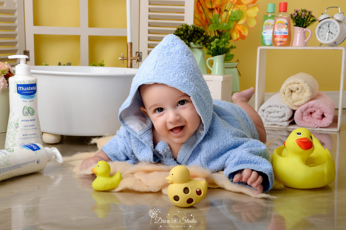 drica studio - ensaio fotográfico - xinguara - bebê vestindo roupão com capuz azul deitado no chão, brincando com patinhos amarelo de borracha - nicho com toalhas rosa, amarela e branca dentro - banheiro branca de frente a janela branca - 