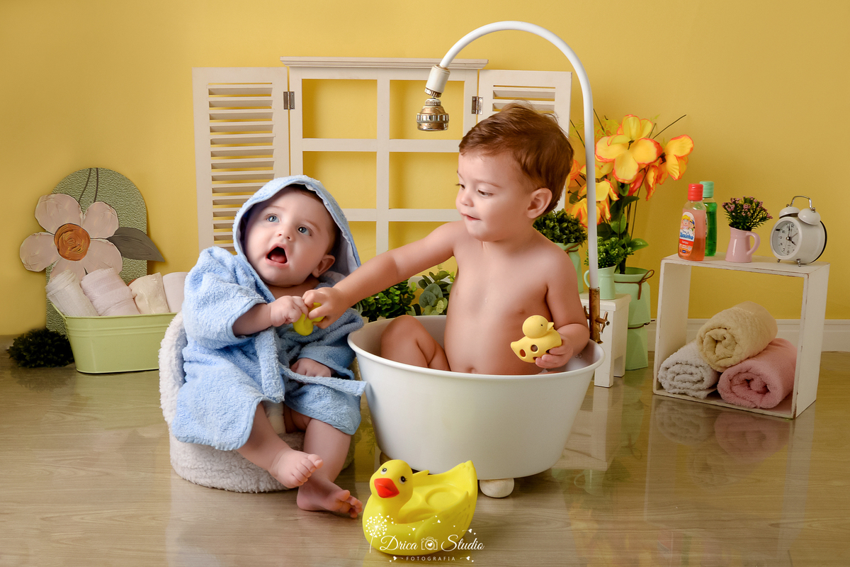 drica studio - ensaio fotográfico - xinguara - dois bebês brincando na banheira - bebê de roupão azul e capuz - menino dentro da banheira com chuveiro segurando patinho -  fundo amarelo