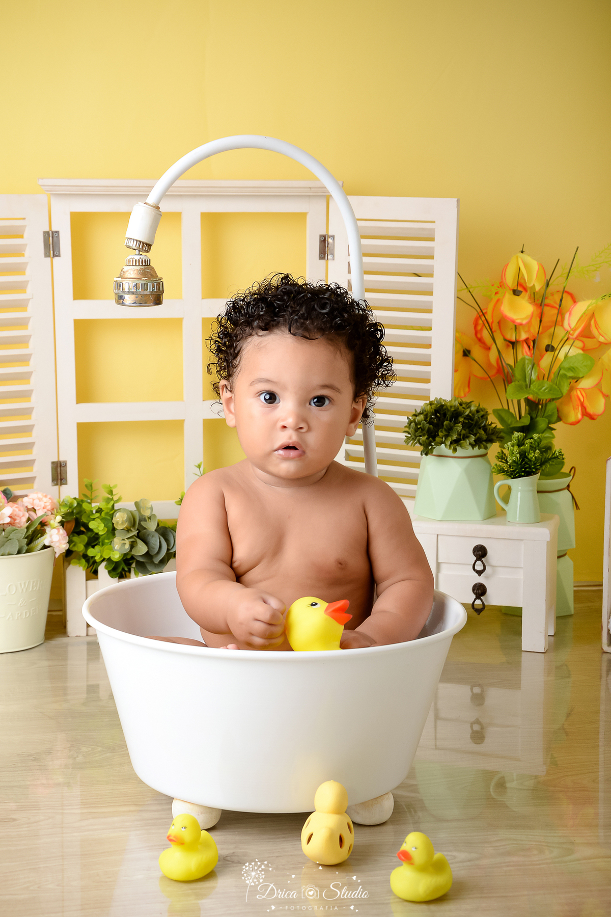 drica studio - ensaio fotográfico - xinguara - menino de cabelos cachiados brincando com patinho de borracha dentro da banheira - cenário com fundo amarelo - cômoda branca com vaso verdes e flores amarelas