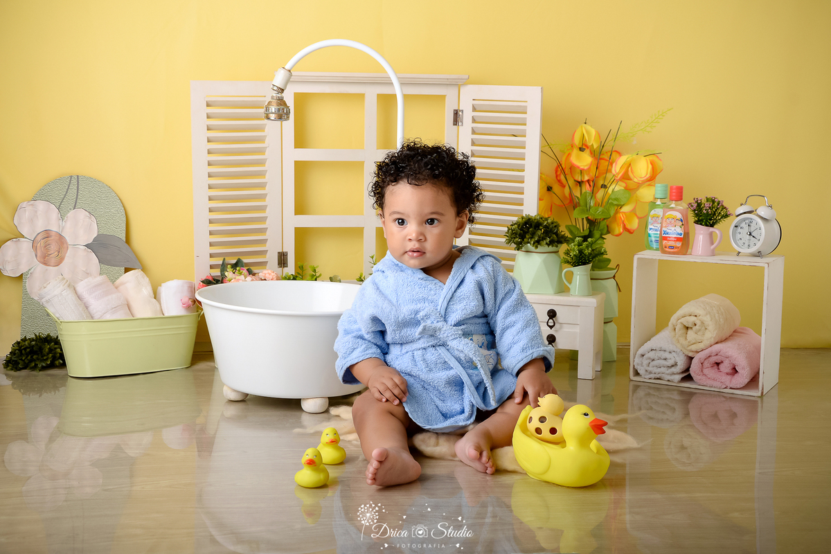 Drica studio - ensaio fotográfico - xinguara - menino de cabelo cacheado vestindo roupão azul- banheira branca - cenário com fundo amarelo - nicho com toalhas dentro - banheira branca com chuveirinho