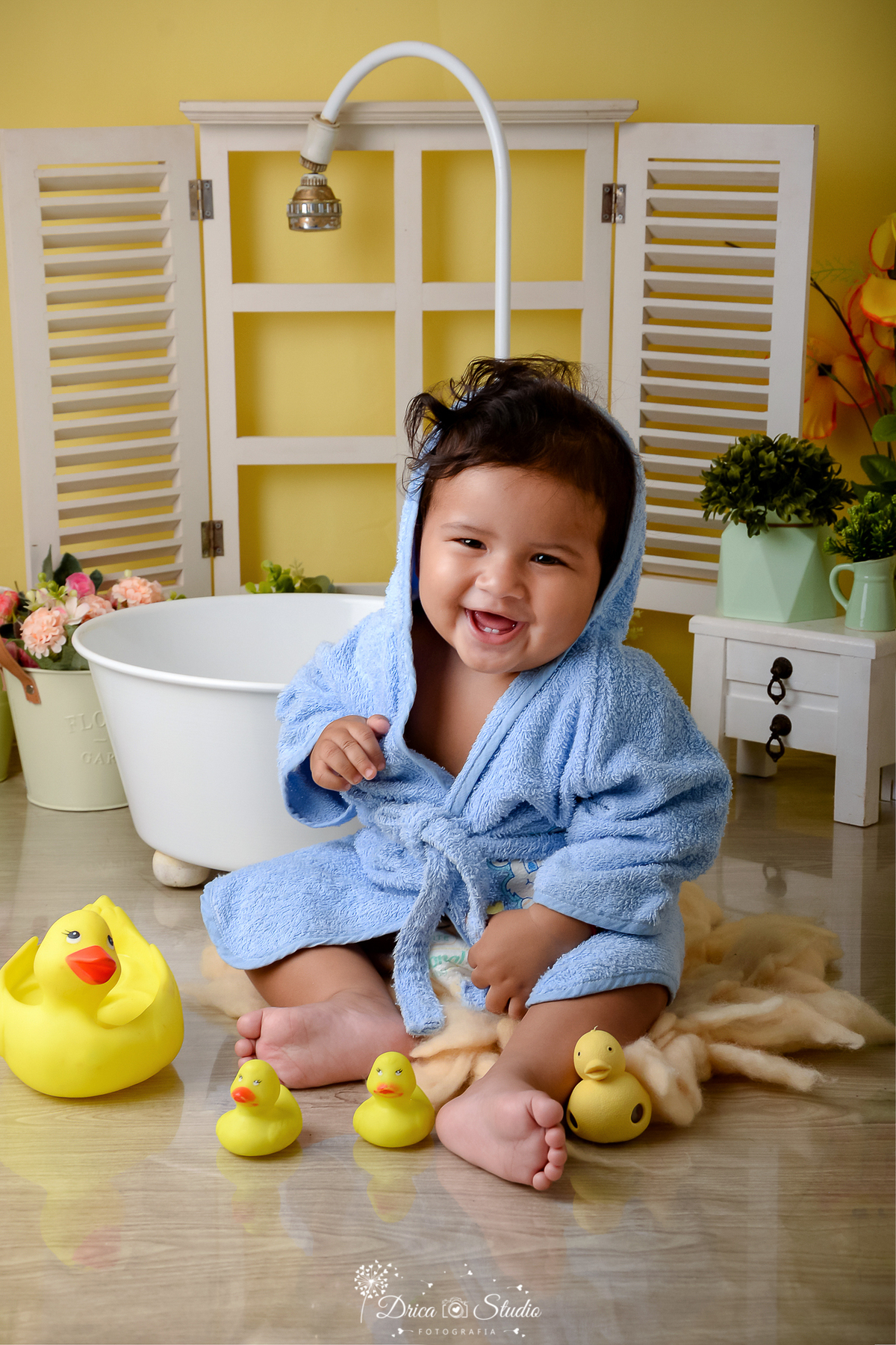 drica studio - ensaio fotografico - xinguara - bebê sorridente vestindo roupão azul claro, sentado ao lada de uma banheira branca - janela branca e cômoda branca ao fundo - cenário fundo amarelo