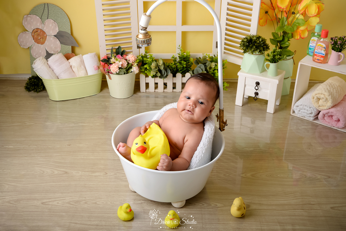 drica studio - ensaio fotográfico - xinguara - bebê banhando em banheira branca com chuveiro, brincando com pato de borracha - patinhos amarelos - cesto verde com toalhas brancas - janela branco com jardineira e arbustos - cômoda branca - funcho amarelo 