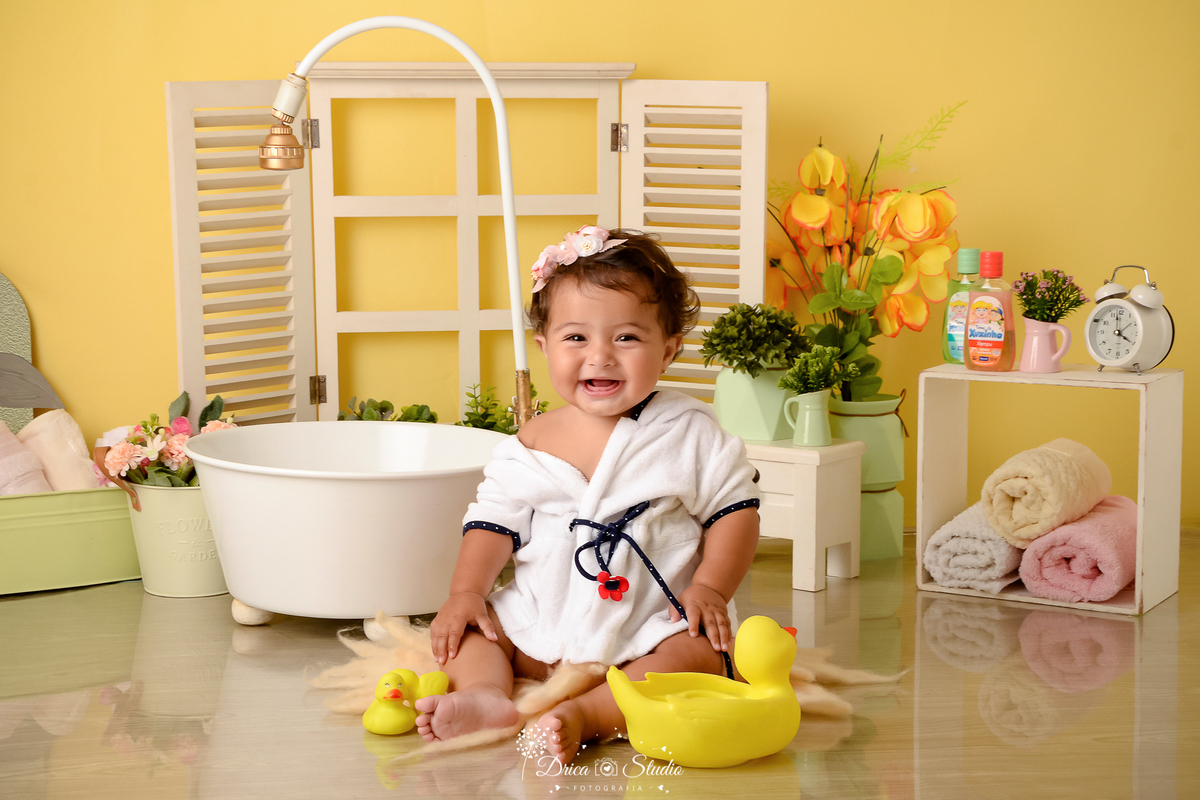 drica studio - ensaio fotográfico - xinguara - menina vestindo roupão branco com detalhe azul, sentada no chão brincando com patinho amarelo de borracha - banheira branca com chuveiro - janela branca com jardineira - nicho com toalhas coloridas 
