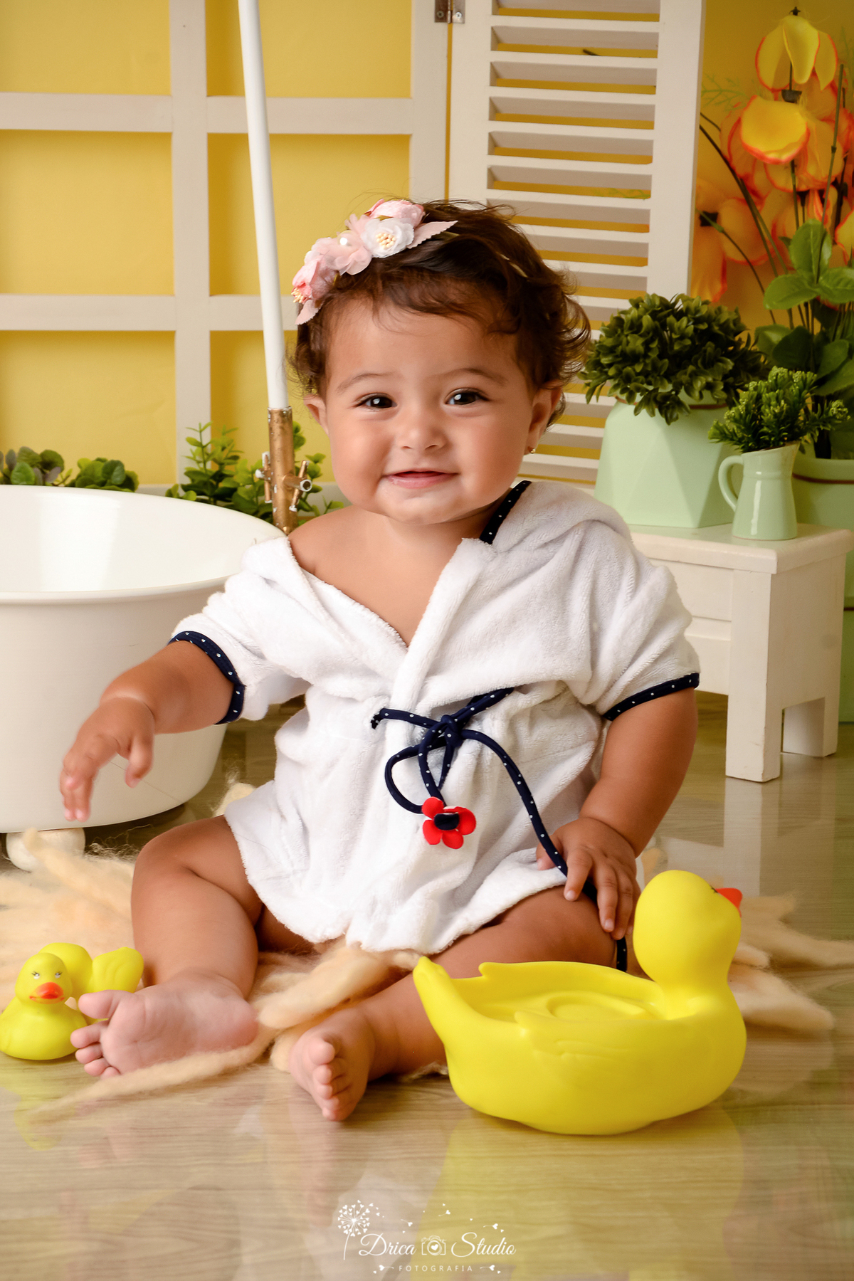 drica studio - ensaio fotográfico - xinguara - menina sorrindo vestindo roupão branco com detalhes azul escuro e uma flozinha na fita - banheira branca - cômoda branca com vasos verdes - fundo amarelo