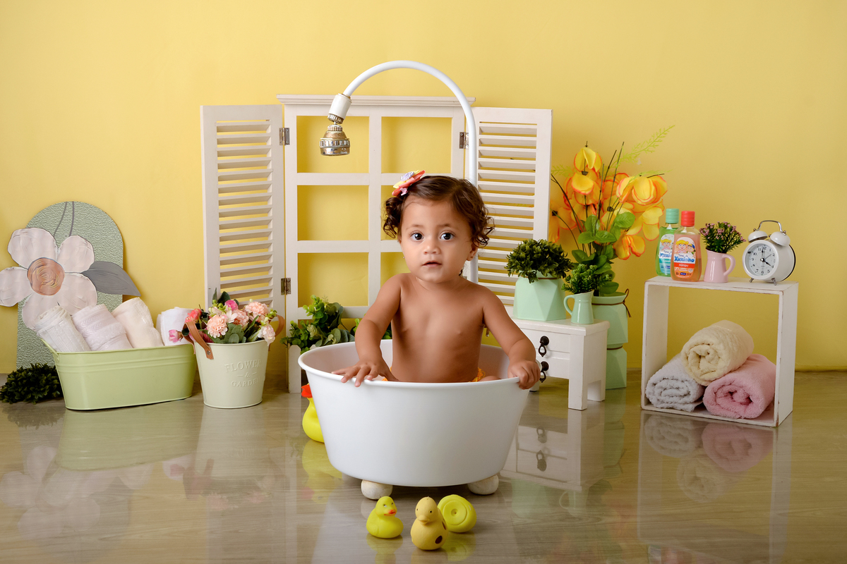drica studio - ensaio fotográfico - xinguara - menina de cabelos cacheados banhando em banheira com chuveiro - balde branco com flores - cesto verde com toalhas brancas - nicho com toalhoas coloridas - flores amarelas - janela branca - fundo amarelo