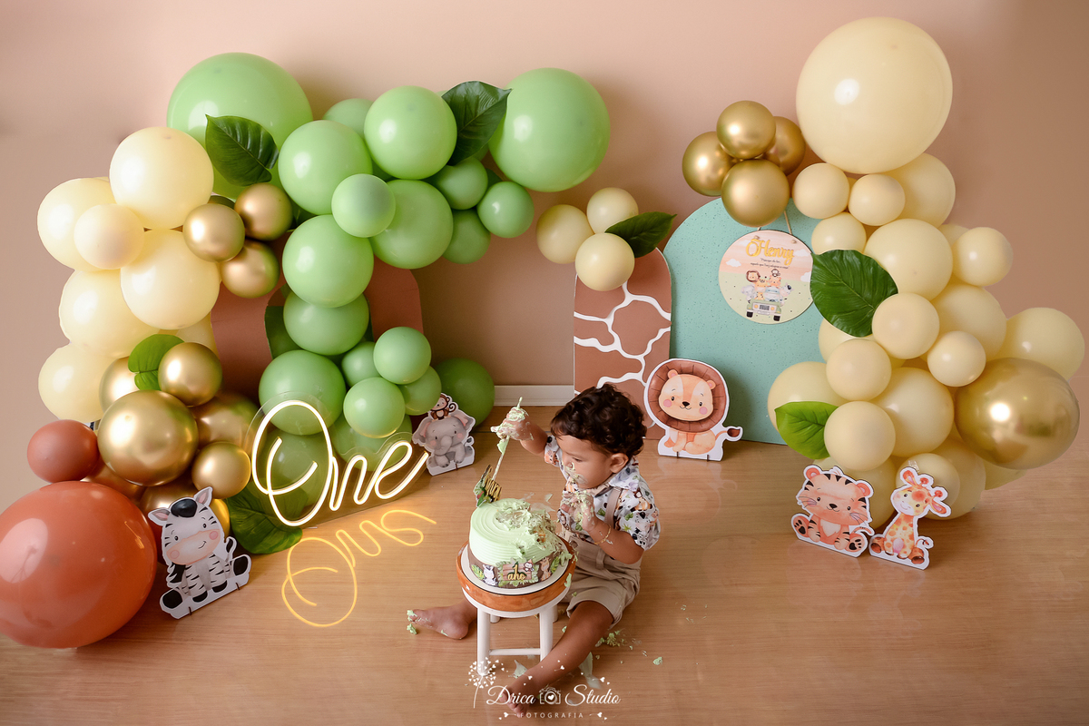drica studio - ensaio fotográfico - xinguara - smahs the cake safari - balões verde, marfim e marrom - menino sentado no chão comendo bolo - one - fundo bege - piso de madeira
