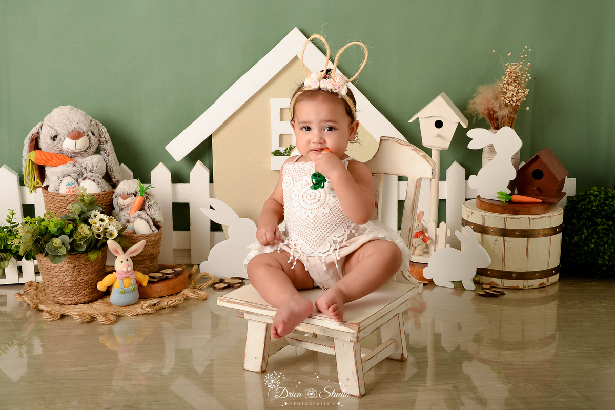 drica studio - ensaio fotográfico infantil - xinguara -  cenario de páscoa - bebe vestindo bory branco sentada em cadeira branca - coelhos - bebe com tiara de orelhas de coelho - fundo verde - cesto de palha - casa de passarinho