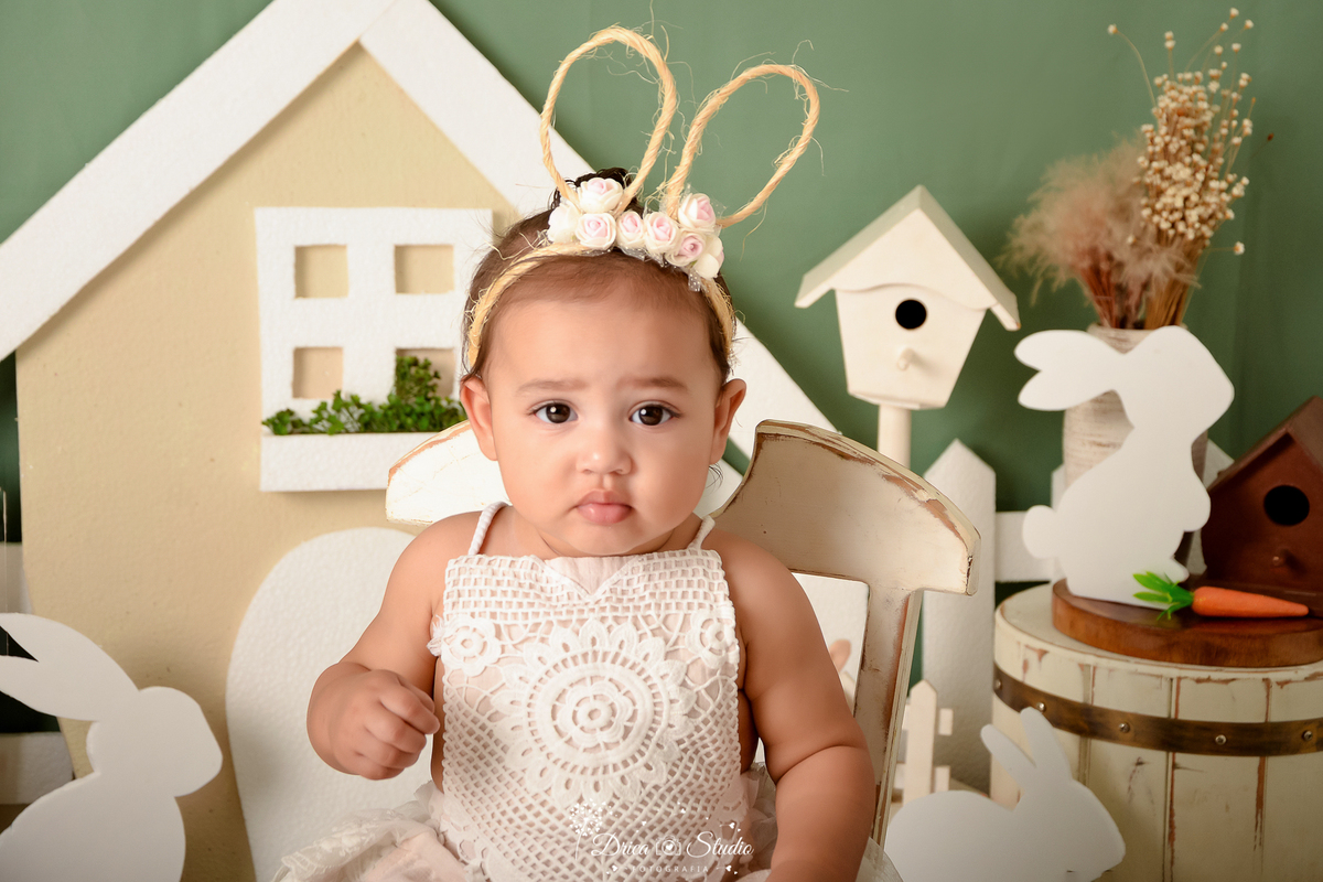 drica studio - ensaio fotográfico infantil - xinguara - cenario de páscoa - fundo verde - casa de coelho - coelho - casa de passarinho - menina vestindo roby de renda - tiara orelhas de coelho - cadeira branca de madeira