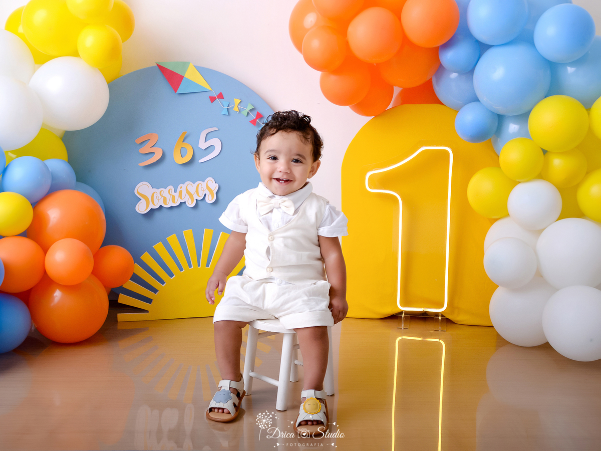 drica studio - ensaio fotográfico - smash the cake - xinguara - 365 sorrisos -balões azul, amarelo, branco e laranja - solzinho - painel azul e amarelo - bebê sorrindo sentado em banquinho de madeira branco
