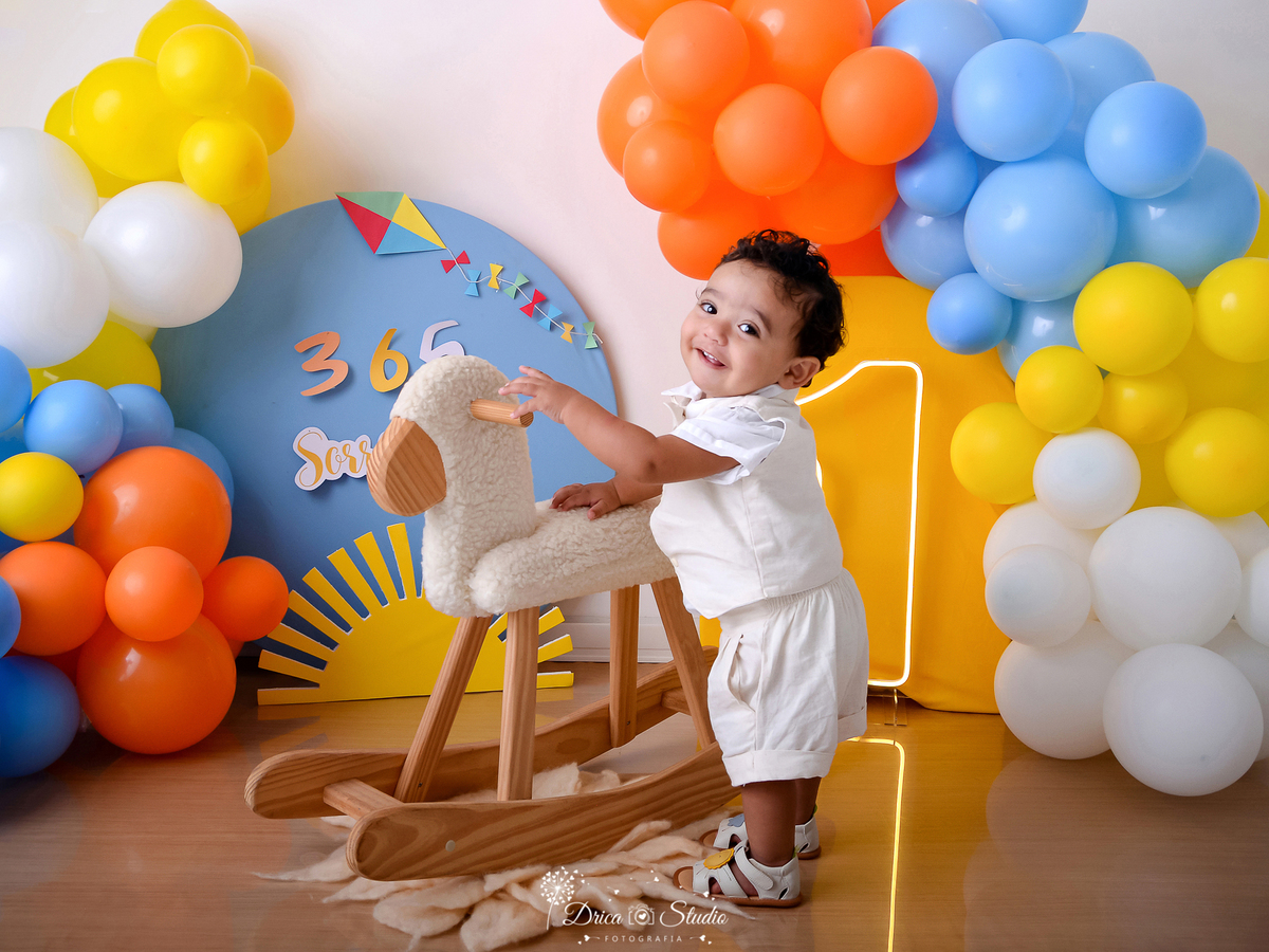drica studio - ensaio fotográfico - smash the cake - xinguara - 365 sorrisos - bebê em pé segurando em ovelha de madeira com acolchoado branco - solzinho - um de led - painel azul e amarelo - 