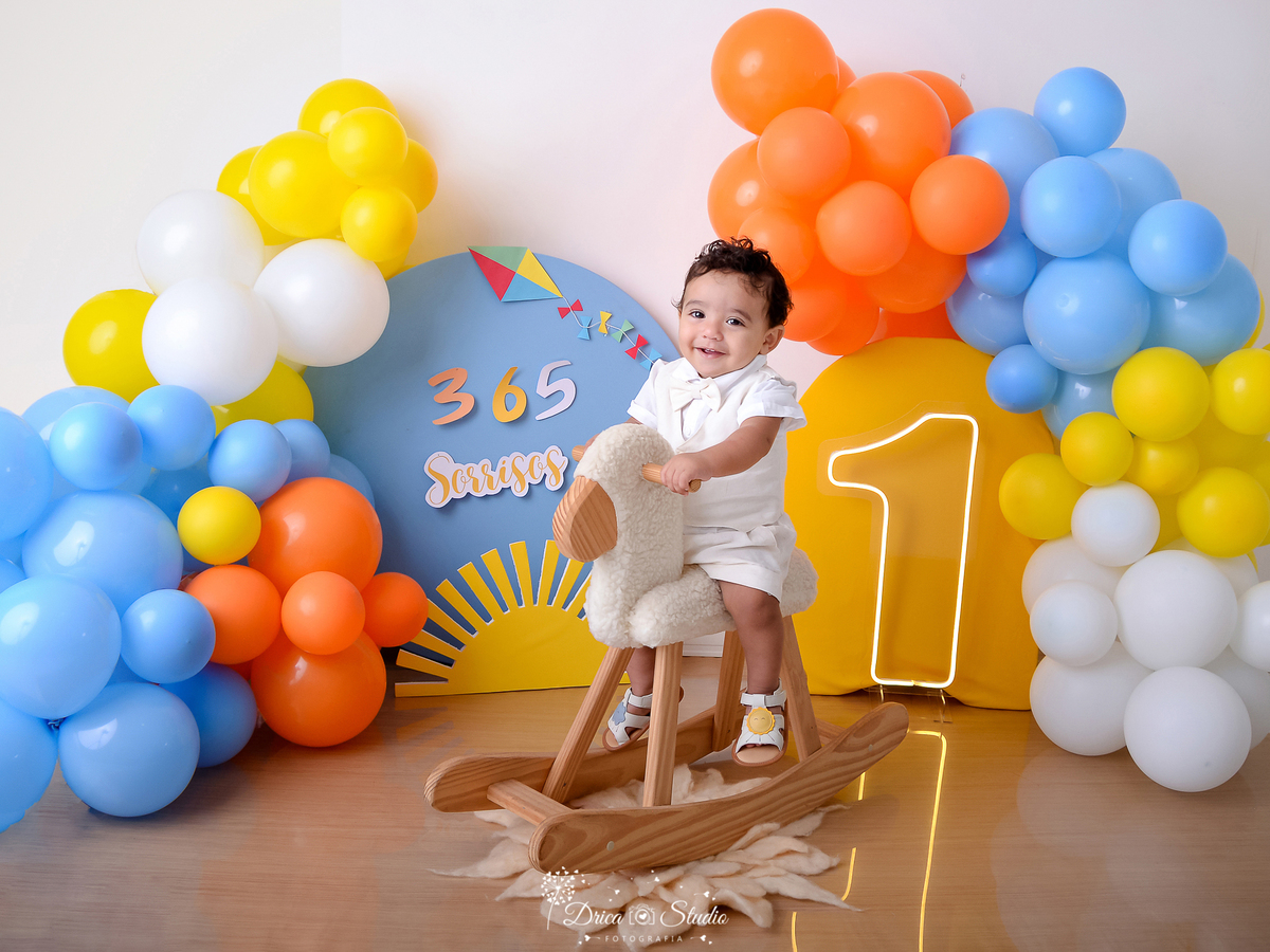 drica studio - ensaio fotográfico - smash the cake - xinguara - 365 sorrisos - bebê montado em cavalinho de madeira - um de led - solzinho - balão azul, amarelo, branco e laranja - painel azul e amarelo - pipa colorida