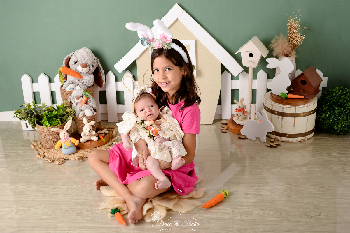 drica studio - ensaio fotográfico infantil - xinguara - páscoa - menina segurando irmã no colo - coelhos - casa de coelho - balde de madeira branco - cesto de palha - fundo verde - tiara de coelho - cenouras