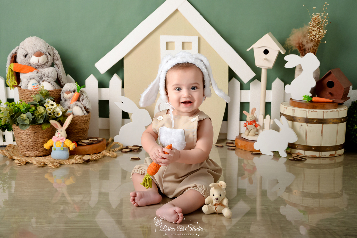 drica studio - ensaio fotográfico infantil - xinguara - páscoa - bebê sentando no chão segurado cenoura - casa de coelho - coelhos - cesto de palha - cesto de madeira branca - piso de madeira - toca com orelha de coelho