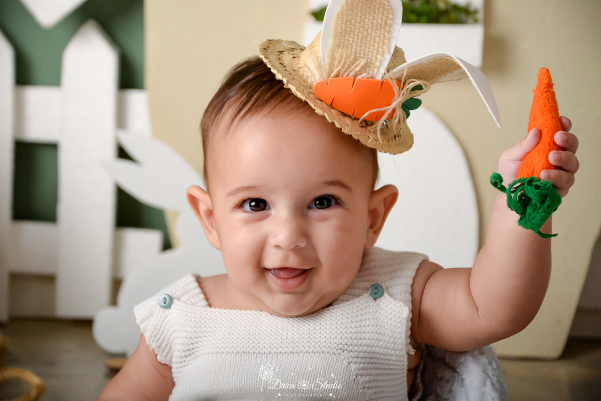 drica studio - ensaio fotográfico infantil - xinguara - páscoa - bebê segurando cenoura - chapéu de palha com orelhas de coelho - casa bege com porta branca - cerca branca - bebê sorridente
