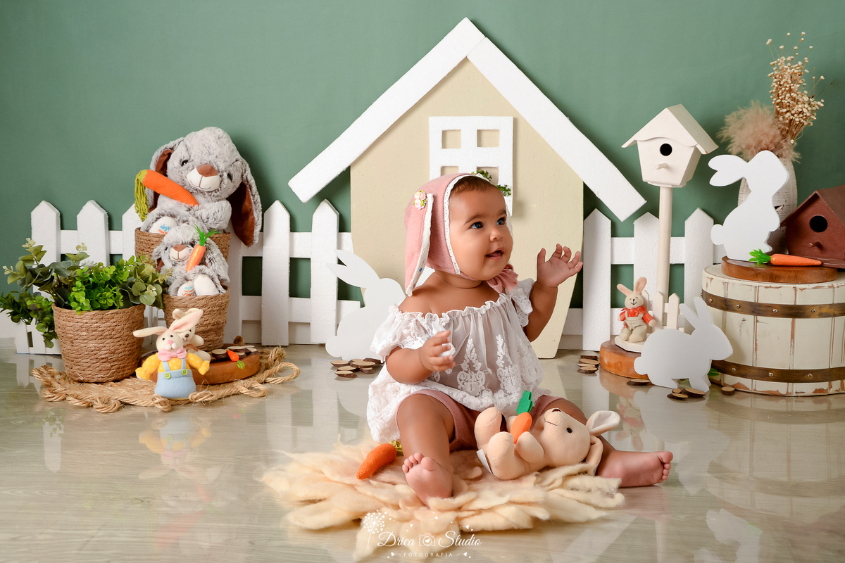 drica studio - ensaio fotográfico infantil - xinguara - páscoa - bebê sentado no chão brincando com coelho de pelucia - coelho - toca de coelho rosa - blusa de renda - casinha com janela branco - cerca branca - cesto de palha