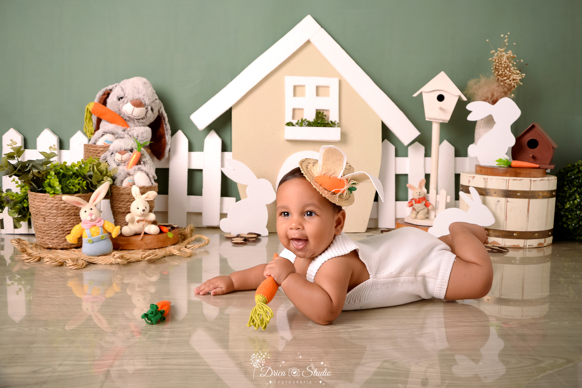 drica studio - ensaio fotográfico infantil - xinguara - páscoa - bebê vestido de coelho deitado no chão de bruços - casinha com porta e janelas branca - coelhos - fundo verde - balde de madeira branco - cesto de palha - cerca branca