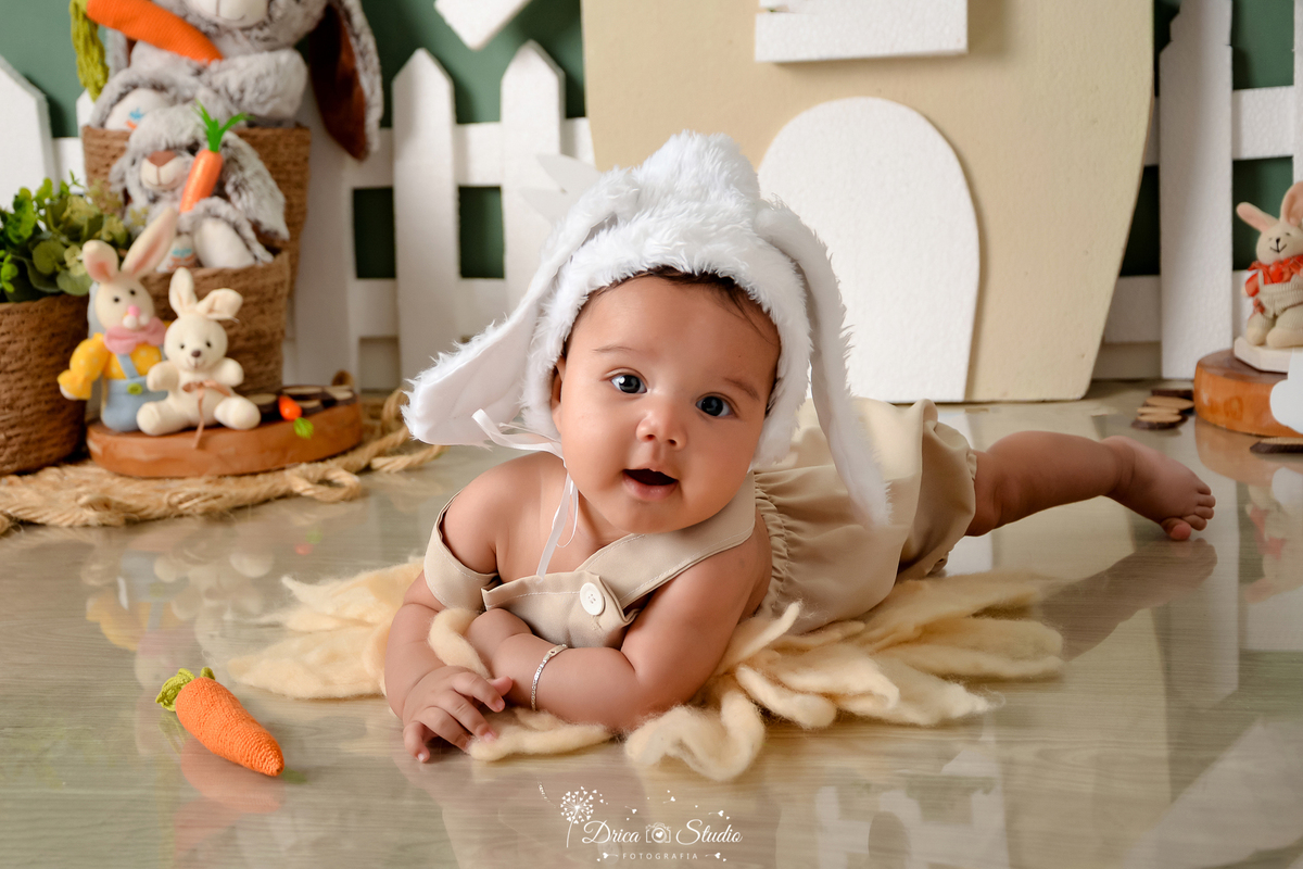 drica studio - ensaio fotográfico infantil - xinguara - páscoa - bebê deitando em tapete felpudo - toca com orelhas de coelho - cenoura - coelho - casinha com janela e porta branca - piso amadeirado - cerca branca