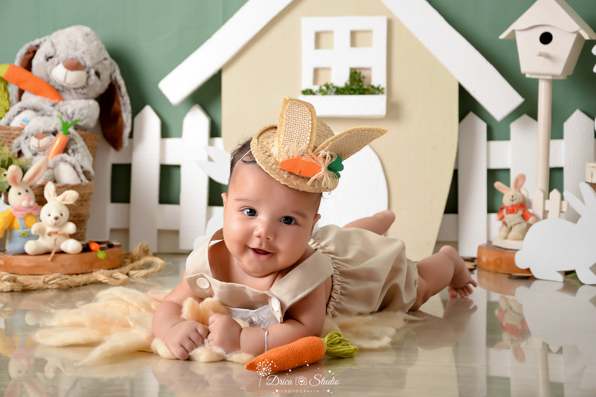 drica studio - ensaio fotográfico infantil - xinguara - páscoa - bebê sorridente deitado de bruços usando chapéu de palha com orelhas de coelho - casinha com portas e janelas brancas - coelhos - cerca branca - fundo verde