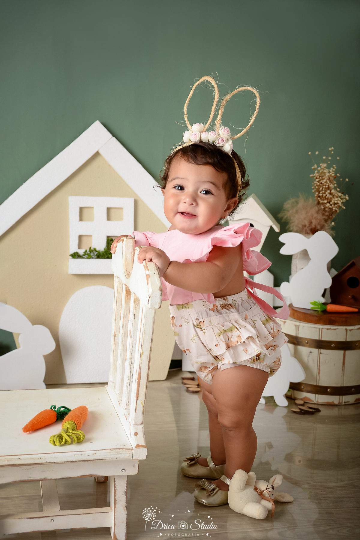 drica studio - ensaio fotográfico infantil - xinguara - páscoa -  bebê em pé segurando no encosto da cadeira - casinha com janela e porta branca - fundo verde - coelho - cesto de madeira branco - ceolho branco