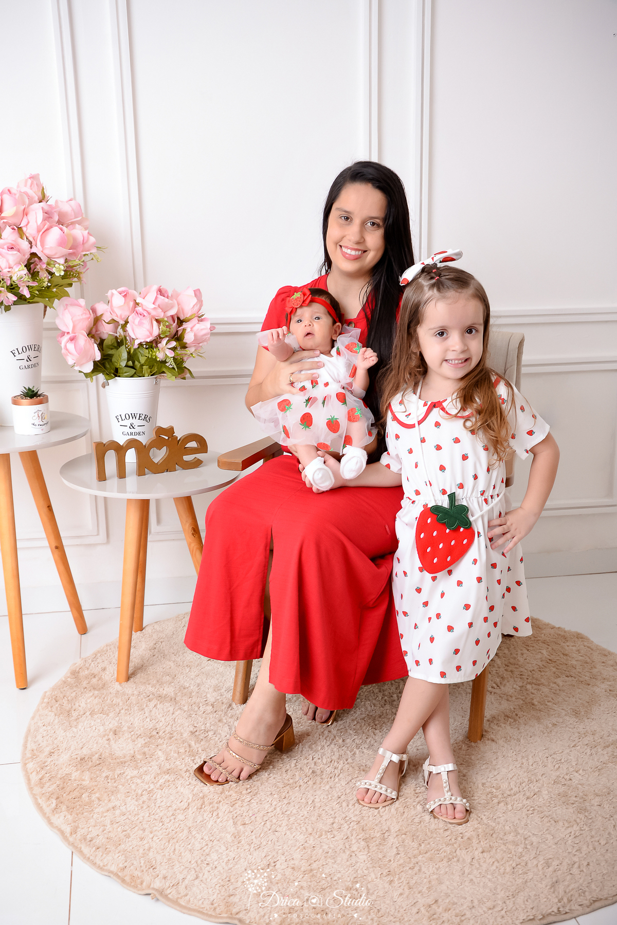 drica studio, ensaio fotografico, xinguara, dia das mães, vestindo branco com moranguinhos, vestindo vermelho, criado mudo, flores rosa, vaso branco, mãe com bebê no colo e filha em pé do lado, boiserie, funco branco