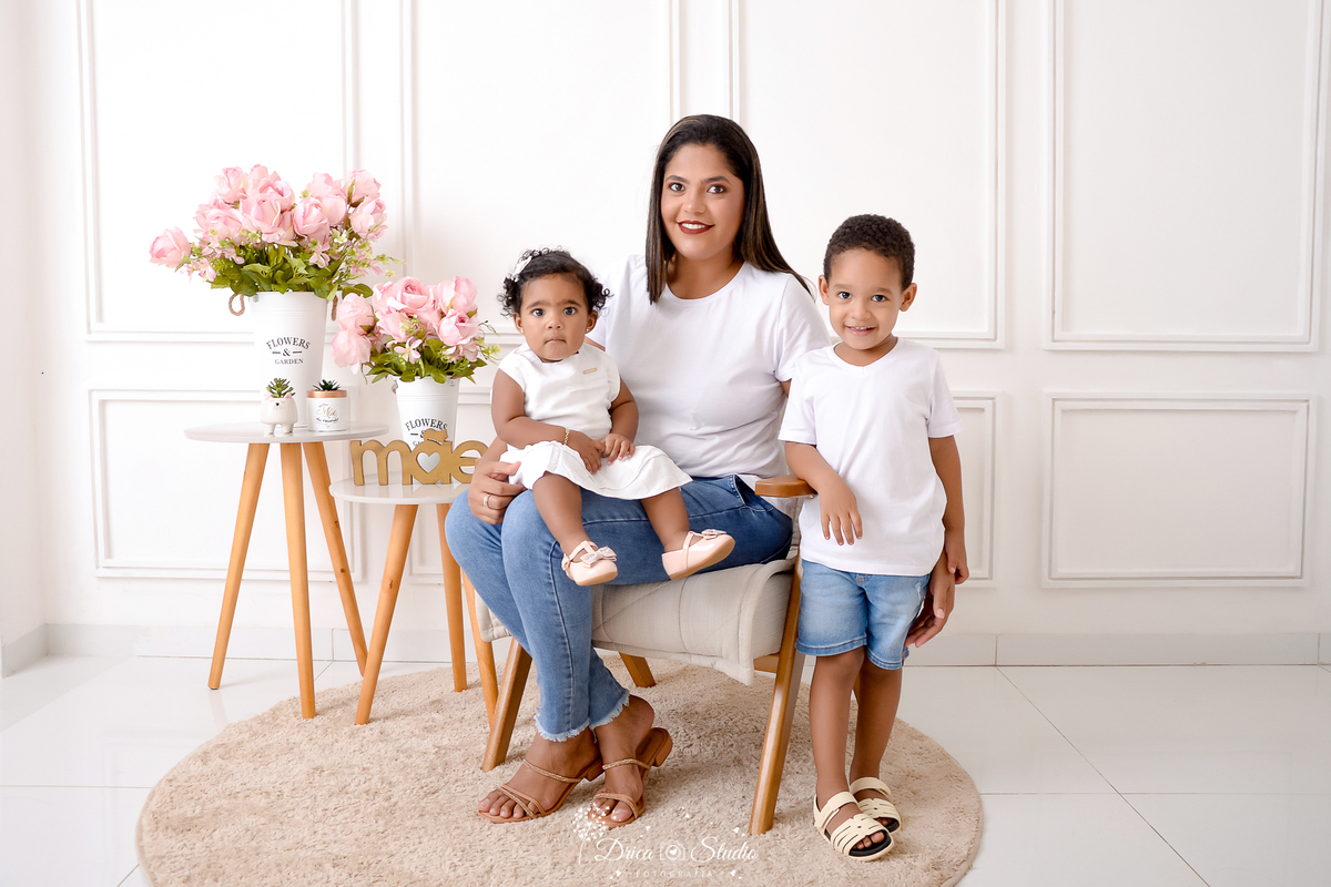 drica studio, ensaio fotografico, xinguara, dia das mães, fundo branco com boiserie, mesas de canto, arrando de flores rosa, vaso branco, tapete redondo berge, mãe com bebê no colo e filho em pé ao lado, camiseta branca, calça jean, short jean 
