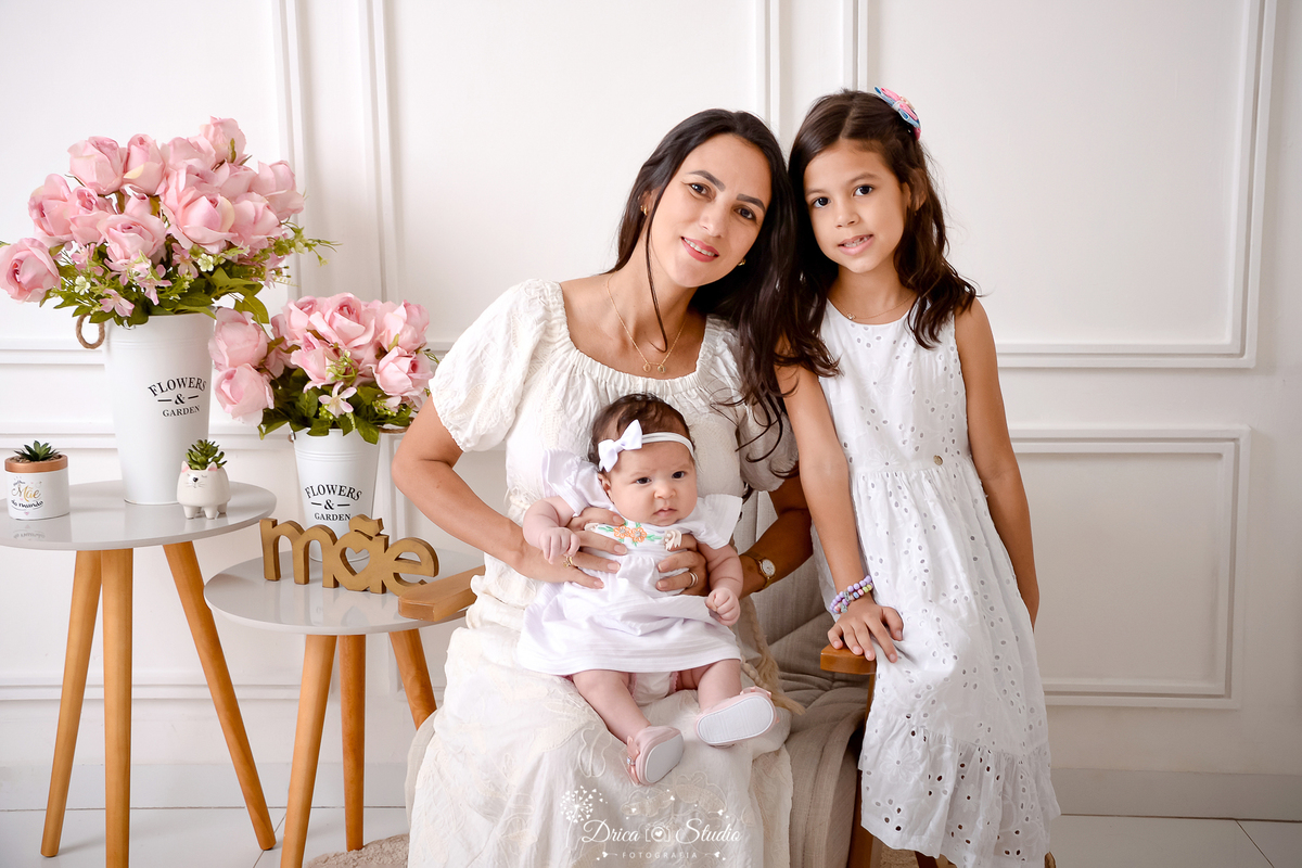 drica studio, ensaio fotografico, xinguara, dia das mães, mãe com bebê no colo e filha em pé ao lado, vestidos brancos, tiara branca, arrundo de flores rosa, mesinhas de canto, fundo branco com boiserie